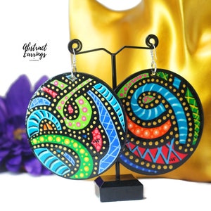 May include: A pair of round, colorful, abstract earrings with a black background. The earrings are painted with bright colors, including blue, green, yellow, orange, and pink. The earrings are hanging from silver hooks.