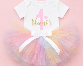 May include: A white baby bodysuit with the number "1" and the name "Eleanor" in gold script, paired with a multi-colored pastel rainbow tutu skirt. The tutu is tied with a white satin bow, creating a festive outfit.