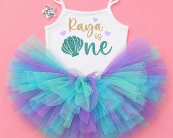 May include: White tank top with the text "Raya is One" in gold glitter and teal. The tank top is paired with a layered tutu skirt in shades of teal, purple, and blue. A silver star is placed on the top left.