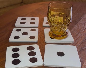 May include: A set of six square white coasters with brown dot patterns, resembling dice. An amber-colored skull-shaped glass sits on one coaster. The coasters are on a wooden table.