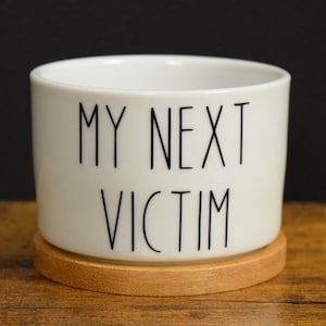 Puede incluir: Maceta de cerámica blanca con base de madera. La maceta tiene texto negro que dice "MY NEXT VICTIM".