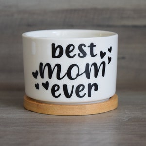 May include: White ceramic planter with a black design that says "best mom ever" with small hearts around the text. The planter has a wooden base.