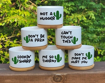 May include: Five white ceramic succulent pots with wooden bases and cactus graphics. The pots are stacked in a pyramid shape and feature humorous sayings such as "Not a hugger", "Don't be a prick", "Can't touch this", "Lookin' sharp", "Me so thorny", and "Don't make me hurt you".