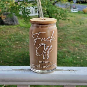 A can shaped glass cup sits on a porch railing. Design on front says Fuck Off I Mean Good Morning in white. Fuck off is in cursive, I mean good morning in a sans serif font. Cup has bamboo lid with glass straight straw.