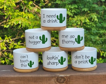 May include: Five white ceramic succulent pots with a green cactus graphic and a funny saying on each. The sayings are "I need a drink", "not a hugger", "me so thorny", "lookin' sharp", and "don't be a prick".