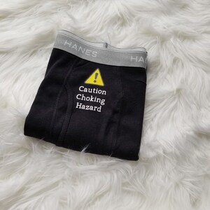 Black boxer briefs lay on a fuzzy white rug. Text on front is caution choking hazard in white with a yellow caution symbol above the text.