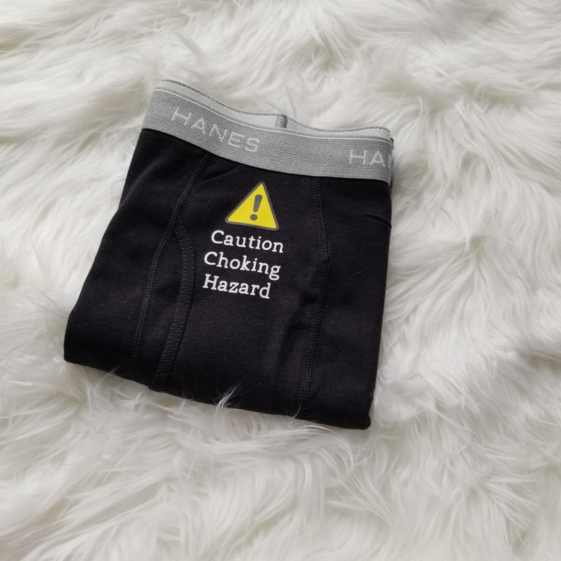 Black boxer briefs lay on a fuzzy white rug. Text on front is caution choking hazard in white with a yellow caution symbol above the text.