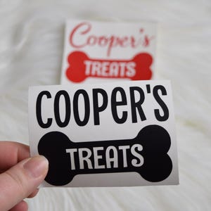 May include: A white sticker with the text "COOPER'S TREATS" in a bold, black font, with the word "TREATS" inside a black bone shape. A second sticker with the same design is in the background, with red text.