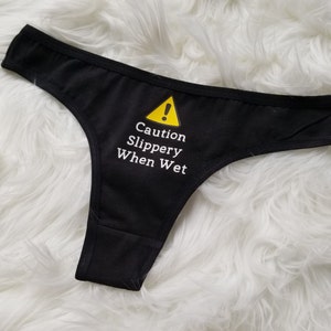 Black thong laying on white fuzzy rug with saying caution slippery when wet in white text. Yellow caution symbol is above the text.