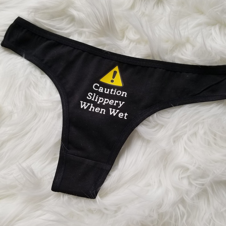 Black thong laying on white fuzzy rug with saying caution slippery when wet in white text. Yellow caution symbol is above the text.