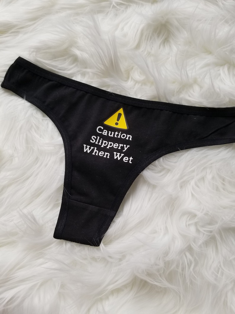 Black thong lays on a white fuzzy rug with text saying Caution Slippery When Wet in white writing with a yellow caution sign above it.