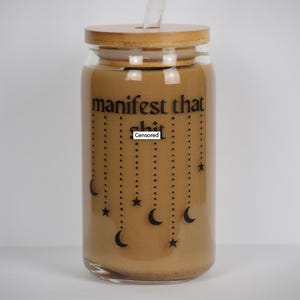 May include: A glass jar with a bamboo lid and a brown liquid inside. The jar has a black design with the text "manifest that [censored]" and stars and crescent moons.