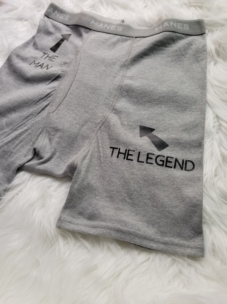 Grey boxer briefs lay flat on a white fuzzy rug. Design says The Man pointing up to the top of the underwear toward the person wearing them, then text saying the legend has an arrow pointing towards the crotch of the underwear.