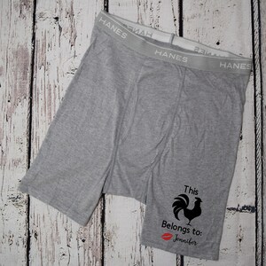 Grey boxer briefs lay flat on a white faux wood backdrop. Design on left leg shows text this cock belongs to, red kissy lips, and the name Jennifer. The cock is not text but an image of a rooster.