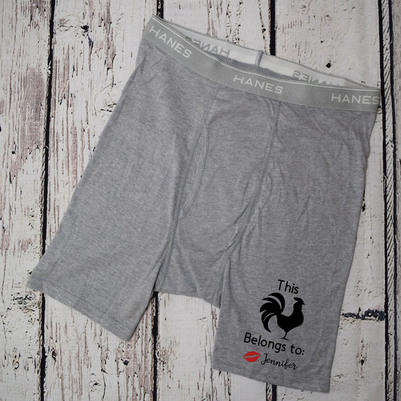 Grey boxer briefs lay flat on a white faux wood backdrop. Design on left leg shows text this cock belongs to, red kissy lips, and the name Jennifer. The cock is not text but an image of a rooster.