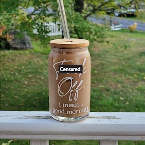 A can shaped glass cup sits on a porch railing. Design on front says Fuck Off I Mean Good Morning in white. Fuck off is in cursive, I mean good morning in a sans serif font. Cup has bamboo lid with glass straight straw.