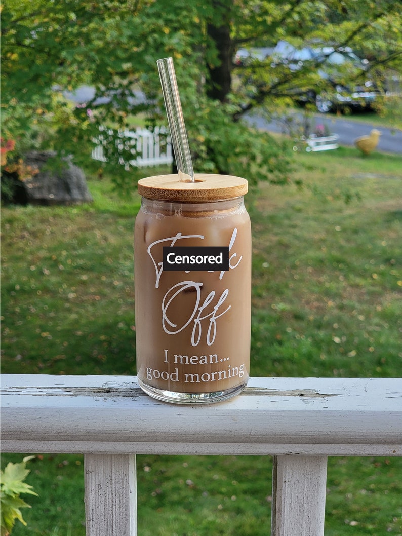 A can shaped glass cup sits on a porch railing. Design on front says Fuck Off I Mean Good Morning in white. Fuck off is in cursive, I mean good morning in a sans serif font. Cup has bamboo lid with glass straight straw.