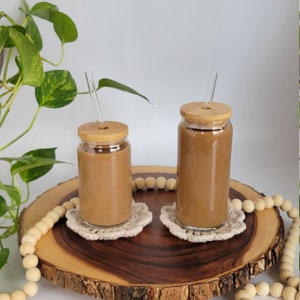 Size difference shown between 16oz and 20oz glasses. Glasses sit on a wooden slab, are filled with coffee, and have bamboo lids and straight glass straws.