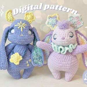 May include: Two handmade crochet bunny dolls in shades of purple and blue. The dolls have long ears, star and flower accents, and the text "Digital pattern" at the top. One doll has a ruffled collar. The dolls are whimsical and soft.