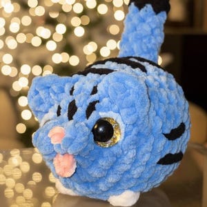 May include: A handmade blue plush cat with black stripes and a black and white tail. The cat has large, glittery gold eyes and a pink tongue sticking out. The cat is sitting on a reflective surface.