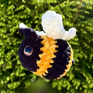 May include: A handmade plush bee with a black and yellow striped body, white wings, and a large, reflective eye. The bee is made of soft, textured yarn and is set against a blurred green background.