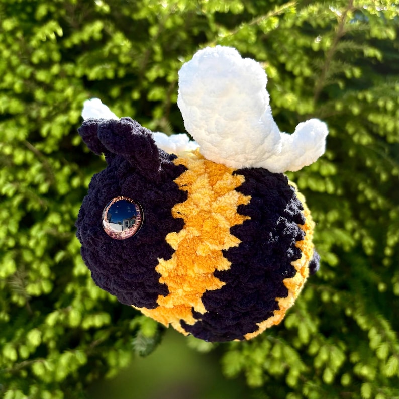 May include: A handmade plush bee with a black and yellow striped body, white wings, and a large, reflective eye. The bee is made of soft, textured yarn and is set against a blurred green background.