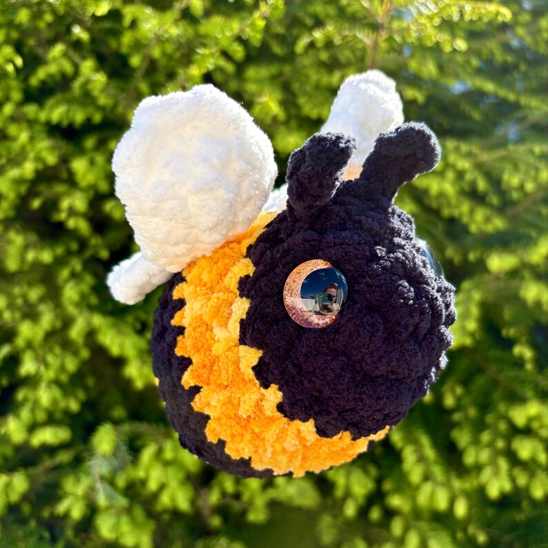 May include: A handmade crochet bee with a black and yellow body, white wings, and large, reflective eyes. The bee is suspended against a backdrop of green foliage, showcasing its soft, textured design. This whimsical item is perfect for home decor or as a unique gift.