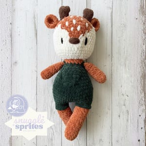 May include: A handmade crochet deer doll with a white head, brown antlers, and orange-brown arms and legs. The deer wears a dark green jumpsuit. The doll has a friendly expression and is part of the Snuggle Sprites Collection.