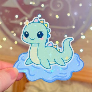 Puede incluir: Una pegatina holográfica con un lindo dinosaurio verde azulado claro con grandes ojos azules, una estrella amarilla y detalles coloridos. El dinosaurio está sobre una nube azul con detalles de estrellas. Acabado brillante e iridiscente.