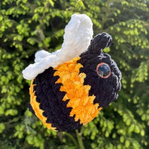 May include: A plush, handmade bee with a black body, yellow and orange stripes, and white wings. The bee has large, reflective eyes. The background is a blurred view of green foliage.