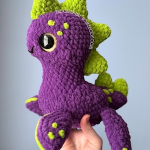 May include: A handmade purple crochet dinosaur plush toy with green detailing. The toy features large black eyes, green spots, and a green spiky dorsal fin. The text "THE CELESTIAL HOMEBODY.COM" is visible on the side.