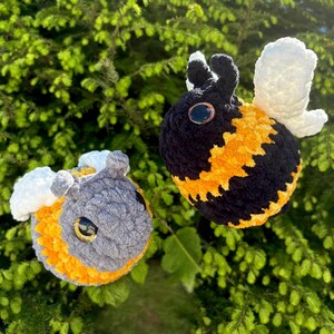 May include: Two handmade crochet bees with black, yellow, and gray bodies, white wings, and large, round eyes. The bees are set against a backdrop of green foliage, creating a whimsical and natural scene.