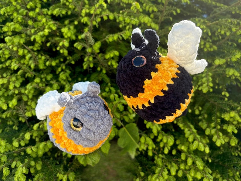 May include: Two handmade crochet bees with black, yellow, and gray bodies, white wings, and large, round eyes. The bees are set against a backdrop of green foliage, creating a whimsical and natural scene.