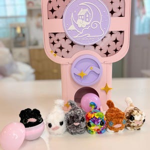 May include: A pink gumball-style machine with a circular emblem featuring a house, moon, and clouds, labeled "CELESTIAL HOMEBOOT". Six small, plush, crocheted animal figures in black, white, gray, rainbow, brown, and spotted colors sit in front.