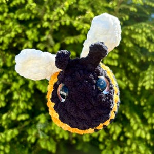 May include: A handmade crochet bee with a black body, yellow stripes, and white wings. The bee has large, reflective eyes and is set against a backdrop of green foliage. This plush toy is a charming decorative item.