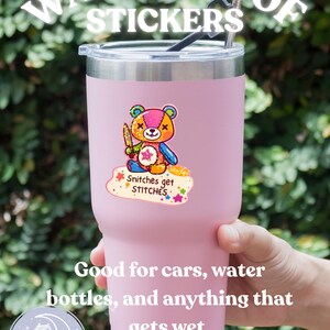K&ouml;nnte beinhalten: Ein rosa isolierter Becher mit Edelstahlrand, mit einem bunten Aufkleber eines Teddyb&auml;ren, der ein Messer h&auml;lt. Der Aufkleber lautet "Snitches get STITCHES". Der Text "WATERPROOF STICKERS" befindet sich &uuml;ber dem Becher.