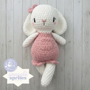 Può includere: Bambola coniglietto bianca all'uncinetto fatta a mano con lunghe orecchie, un fiocco rosa e un vestito rosa. Il coniglietto ha occhi neri, guance rosa e un naso rosa. Parte della collezione Snuggle Sprites.