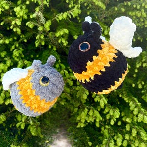 May include: Two handmade crochet bee plushies. One bee is black and yellow, the other is gray and yellow. Both have white wings and large, round eyes. The bees are set against a backdrop of green foliage.