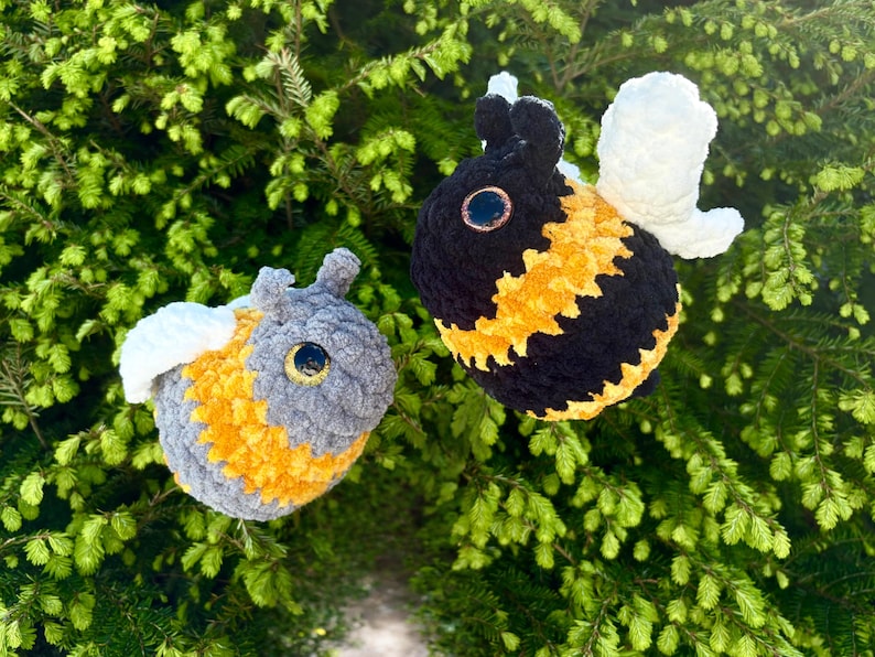 May include: Two handmade crochet bee plushies. One bee is black and yellow, the other is gray and yellow. Both have white wings and large, round eyes. The bees are set against a backdrop of green foliage.