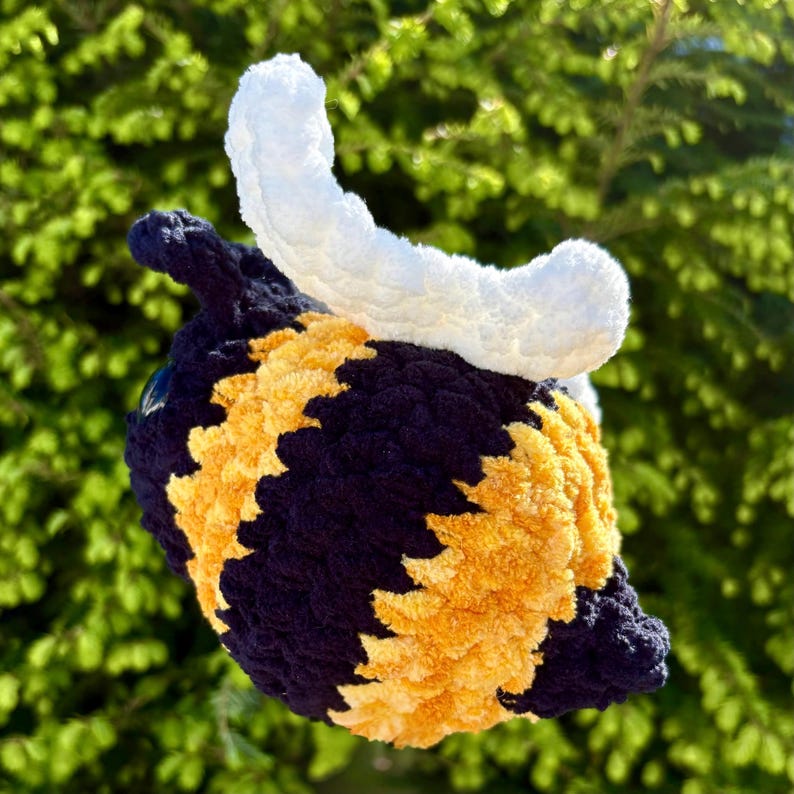 May include: A handmade crochet bee hat with a white horn-like detail. The hat features a navy blue base with yellow and orange stripes. The hat is designed to be worn on the head.