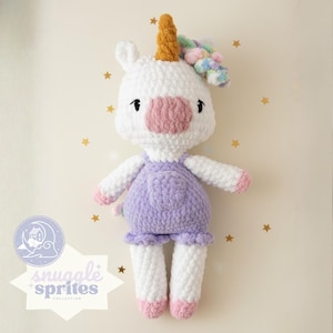 Puede incluir: Muñeca de unicornio de ganchillo hecha a mano con cuerpo blanco, vestido lavanda y detalles rosas. El unicornio tiene un cuerno dorado y una melena colorida. La muñeca es parte de la colección Snuggle Sprites.