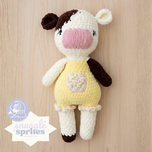 Könnte beinhalten: Handgefertigte Häkelkuhpuppe mit weiß-braunem Kopf, rosa Nase und gelber Latzhose. Die Puppe hat weiße Arme und Beine mit braunen Hufen. Die Latzhose hat eine Blumentasche und rosa-grüne Verzierungen. Teil der Snuggle Sprites Kollektion.