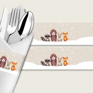 May include: A white napkin with a brown and white patterned napkin ring featuring a raccoon, bear, bunny, and fox in a snowy scene. The napkin ring is wrapped around a knife, fork, and spoon.