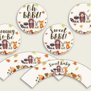May include: Set of cupcake wrappers and toppers featuring a woodland animal theme. The wrappers have a white background with brown, orange, and green leaves and acorns. The toppers feature a raccoon, bear, bunny, and fox with the text "Sweet Baby" and "Mommy To Be".