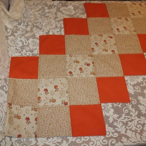 May include: A patchwork table runner with alternating squares of orange and beige fabric. The beige squares feature a floral pattern and a pumpkin pattern.