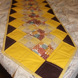 May include: A yellow and brown quilted table runner with a geometric pattern. The runner is made of fabric with a floral print and a honeycomb pattern.