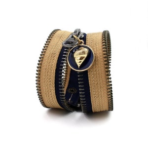 May include: A double zipper bracelet with a gold and blue charm. The bracelet is made of two layers of zipper tape, one beige and one navy blue.