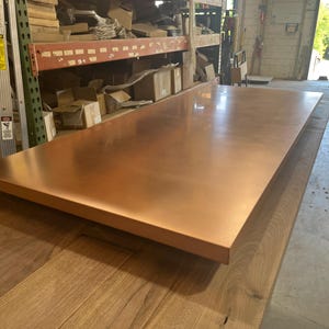 May include: A large, rectangular copper tabletop with a polished, reflective surface. The copper has a warm, reddish-brown hue. The tabletop is resting on a wooden surface, showcasing the contrast between the two materials. The overall aesthetic is industrial and modern.