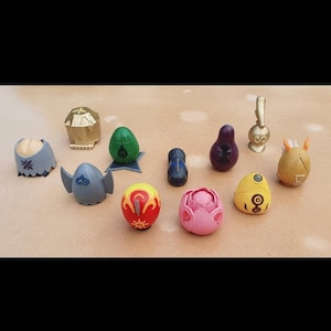 May include: A collection of colorful, stylized egg-shaped figurines. The figurines are in various colors, including gold, green, purple, yellow, and pink. Some have intricate designs and patterns, while others have simple shapes. The figurines are arranged on a light brown surface.
