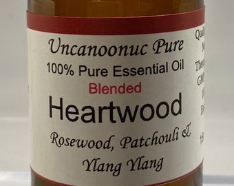 May include: A brown glass bottle with a black cap, labeled "Heartwood" essential oil. The label includes the words "Rosewood, Patchouli & Ylang Ylang". The bottle contains a clear, amber-colored liquid.